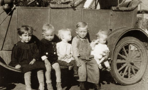An old image of 5 young children sitting on the side of an old vehicle