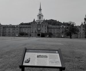 Image of a very large old lunatic asylum. There is a clocktower in the center of the building and then the building stretches out to the edges of the image on both sides.