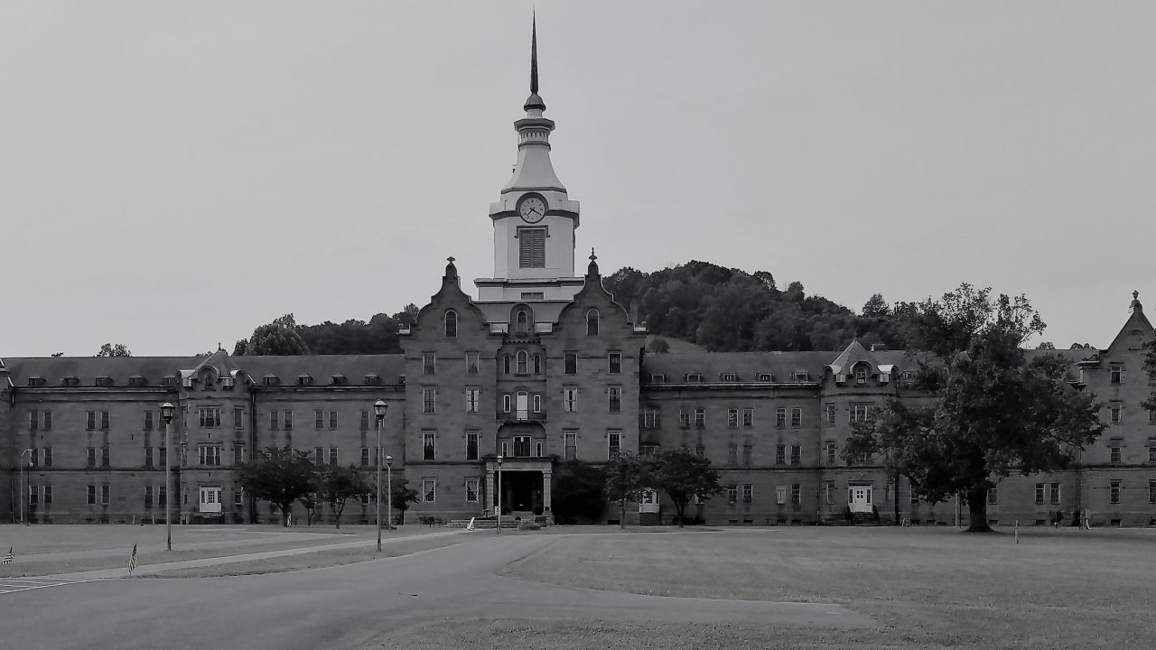 Image of a very large old lunatic asylum. There is a clocktower in the center of the building and then the building stretches out to the edges of the image on both sides.